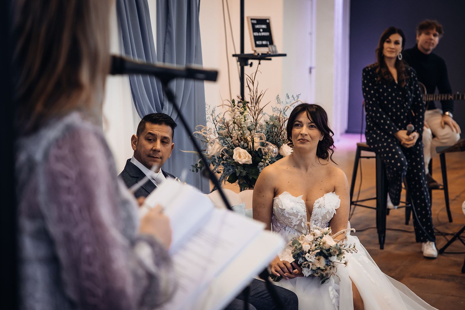 Bruid luistert aandachtig naar de ceremoniespreker tijdens een persoonlijke huwelijksceremonie, omringd door dierbaren in een warme setting.