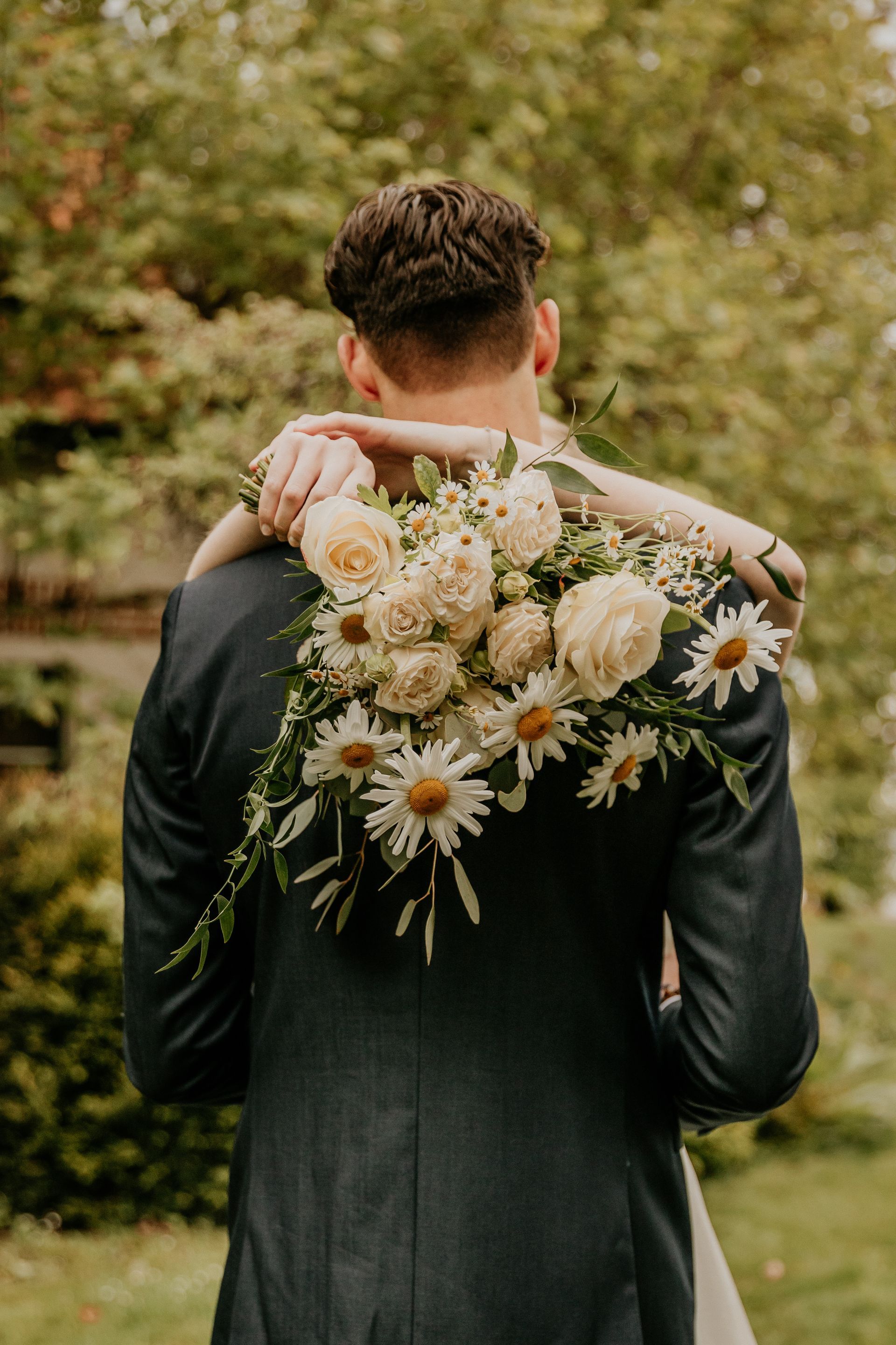 Intiem moment van het bruidspaar omarmd in de natuur, met een bruidsboeket vol zachte bloemen op de achtergrond.