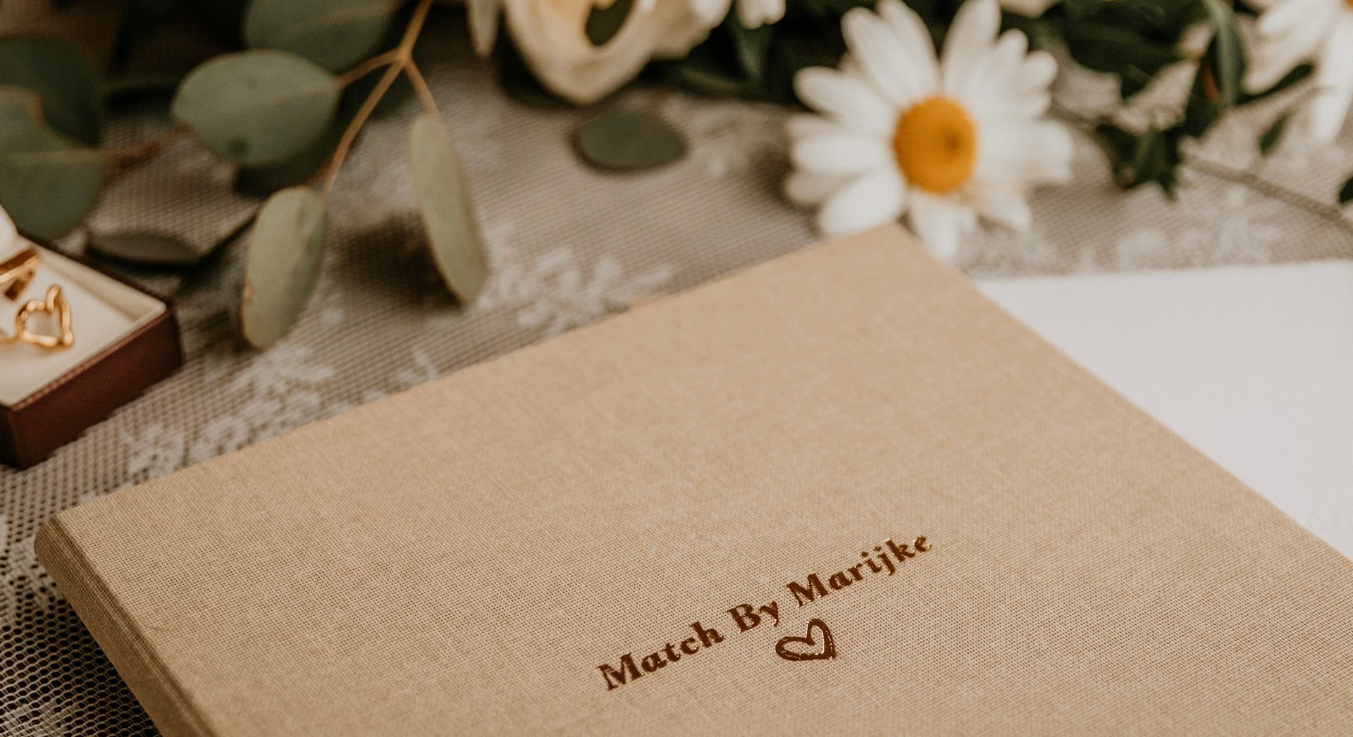 Linnen huwelijksboek met de tekst “Match by Marijke” en een subtiel hartje, detail van een persoonlijke trouwceremonie.