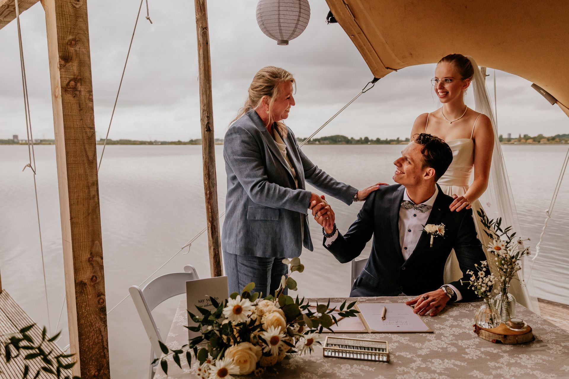 Trouwambtenaar feliciteert het bruidspaar na het ja-woord tijdens een huwelijksceremonie aan het water, in een persoonlijke en warme setting.