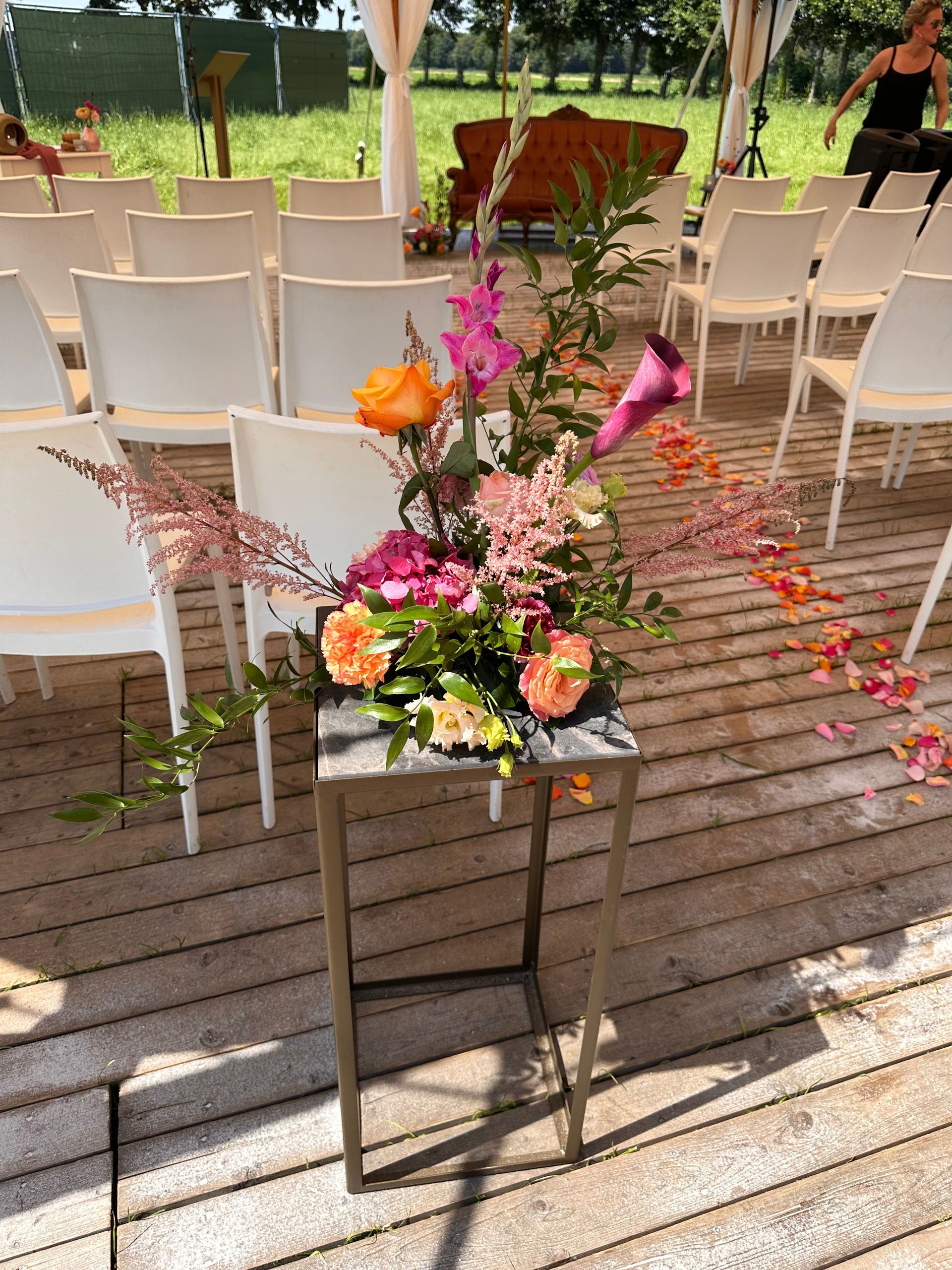 Trouwceremonie-opstelling met witte stoelen, kleurrijk bloemstuk en bloemblaadjes op het gangpad voor een sfeervolle buitenbruiloft.