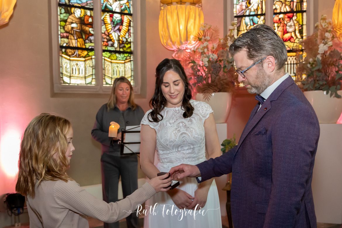 Bruidspaar wisselt ringen uit tijdens een huwelijksceremonie in een kerk, met hulp van een kind en warme, intieme sfeer.