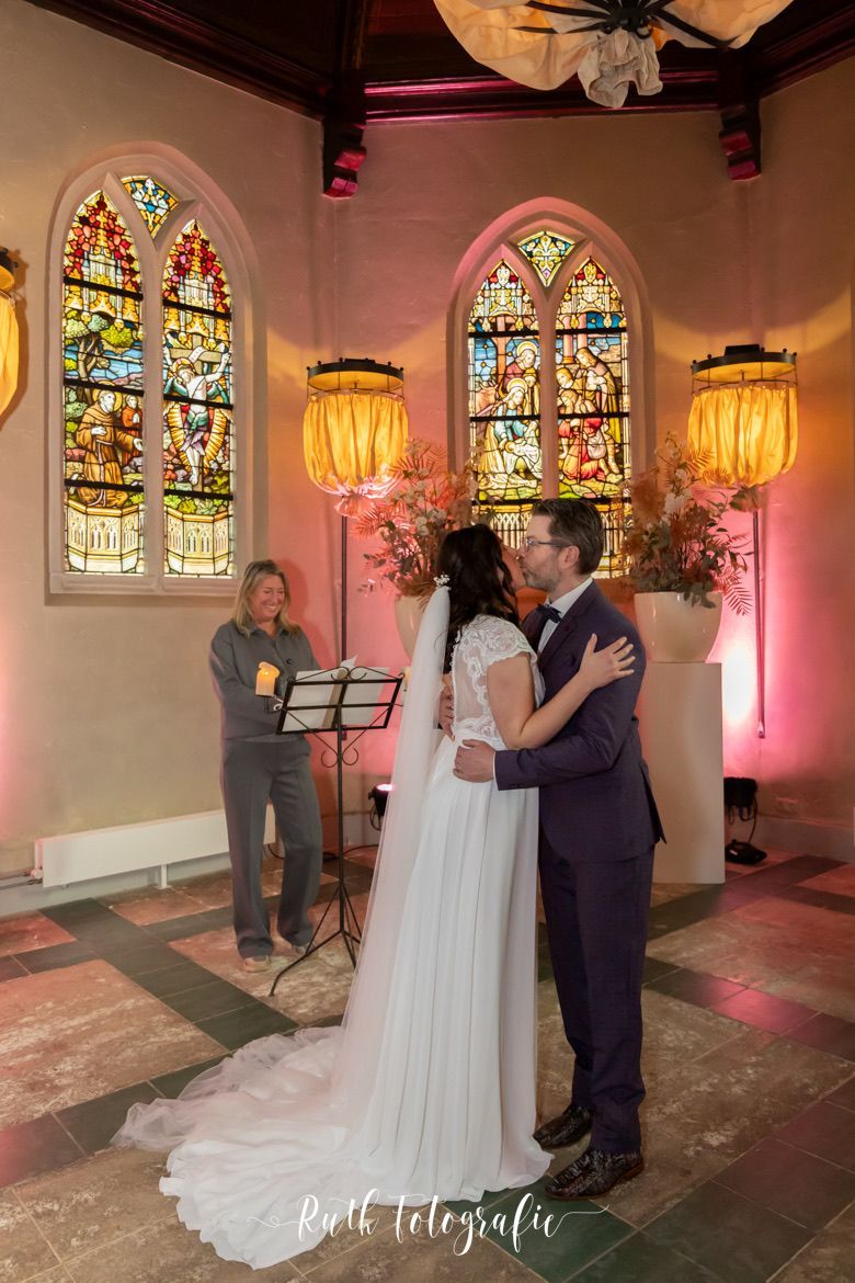 Bruidspaar kust elkaar tijdens een romantische huwelijksceremonie in een kerk met kleurrijke glas-in-loodramen en warme verlichting.