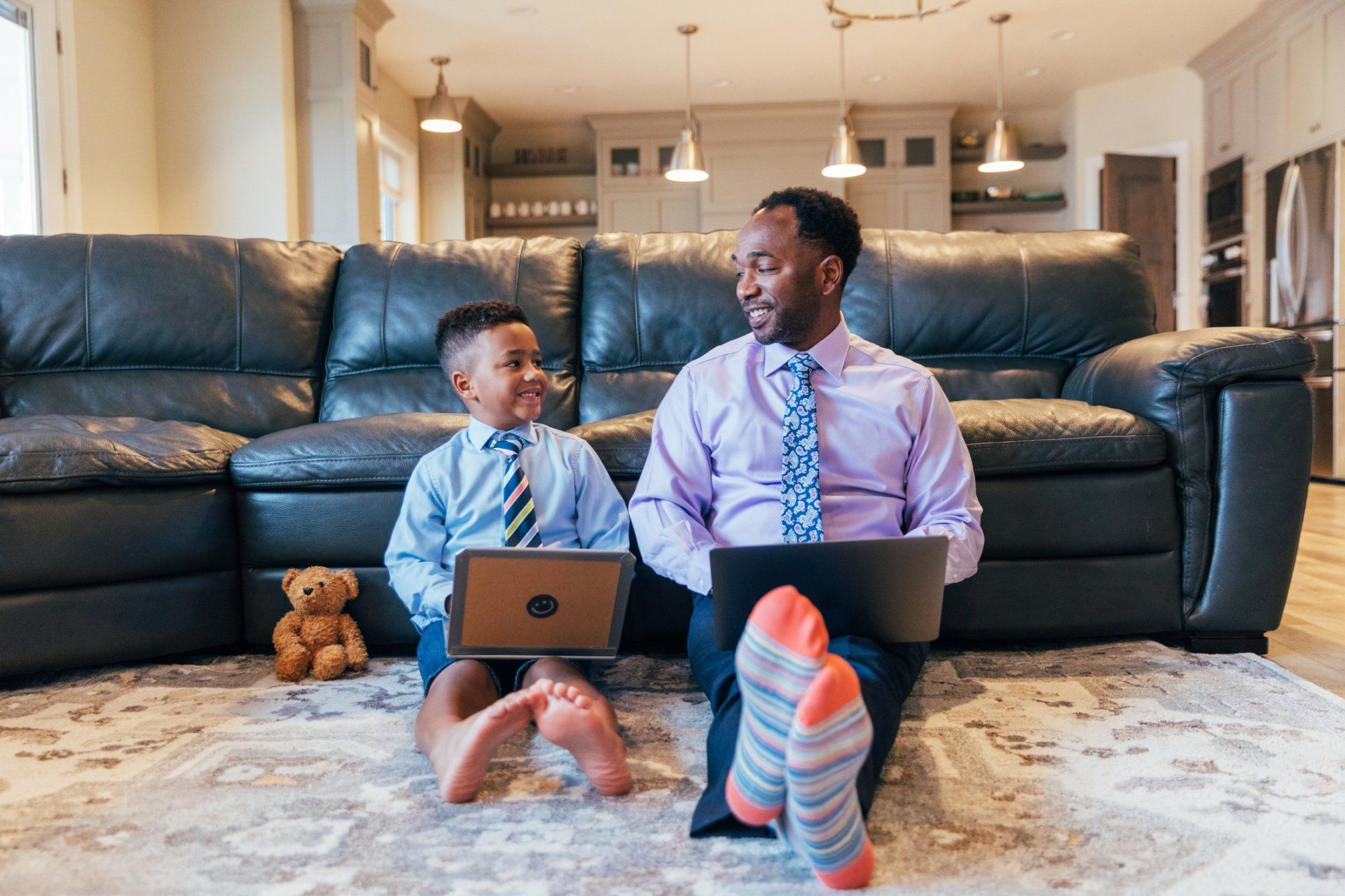 Father and son wearing dress shirts and ties, sitting on the floor with laptops, smiling at each other.