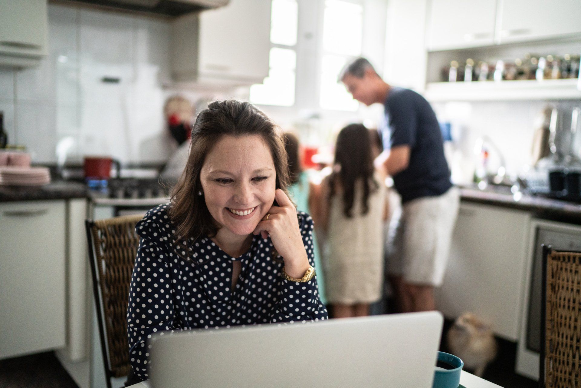 Woman smiling at laptop in kitchen while family preps food.
