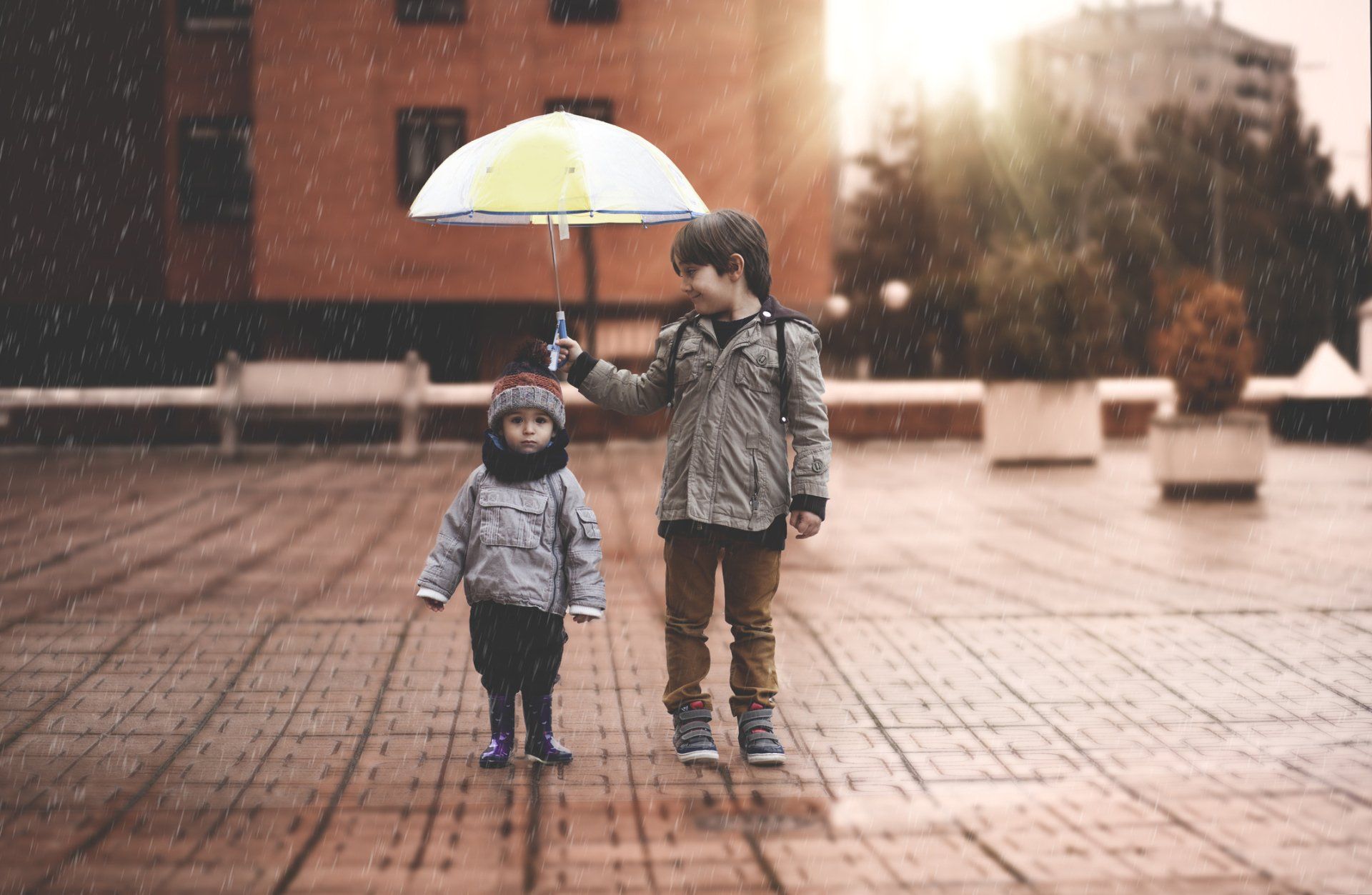 Two children walking in the rain, sharing an umbrella. One child is holding the umbrella, the other is small and looks cold.