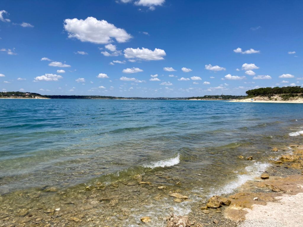 Rocky beach with clear blue water, small waves, and a sunny sky with scattered clouds.