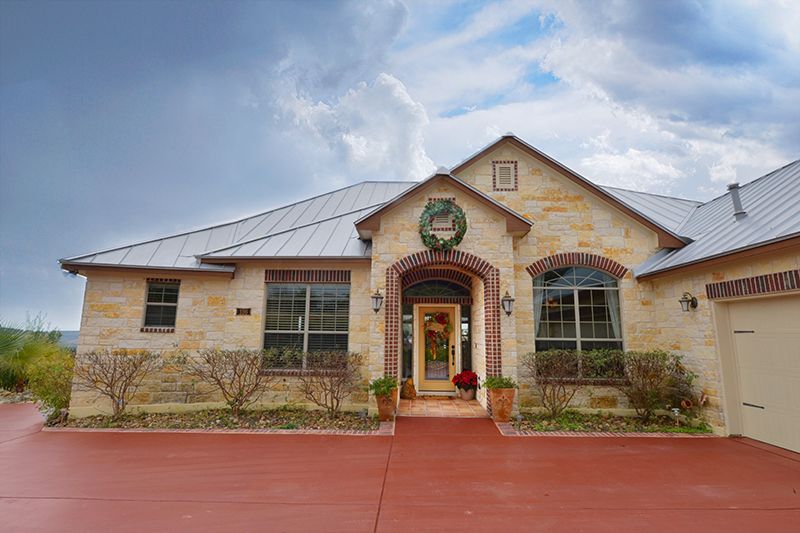 Single-story stone house with a red driveway, arched front entry, and desert landscaping under a cloudy sky