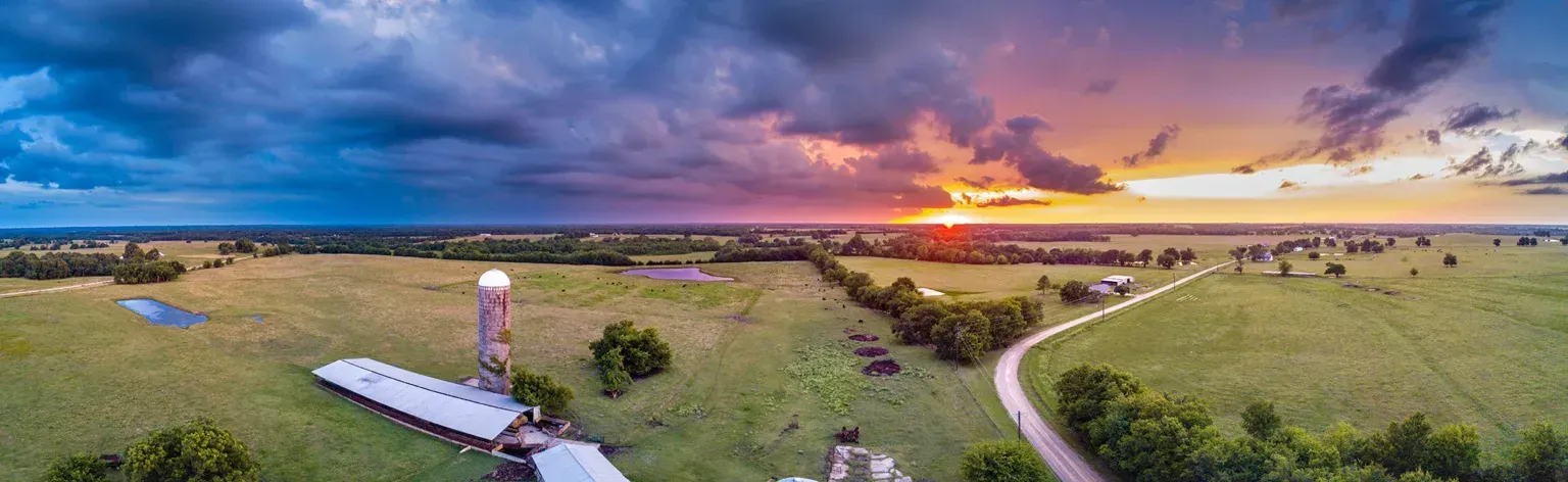 Sunset over a grassy landscape with a winding dirt path, a small tower, and a few buildings in the distance.