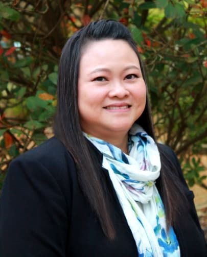 Woman with long dark hair, wearing a black blazer and patterned scarf, smiles outdoors.