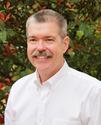 Man with a mustache smiles at the camera, wearing a white button-down shirt, outdoors.