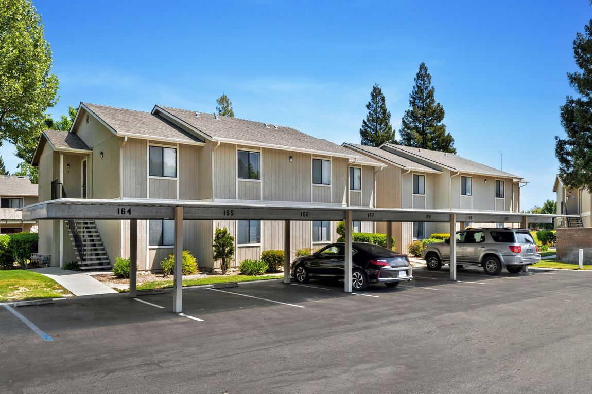 Apartment complex with covered parking; beige siding, blue sky, cars parked beneath.