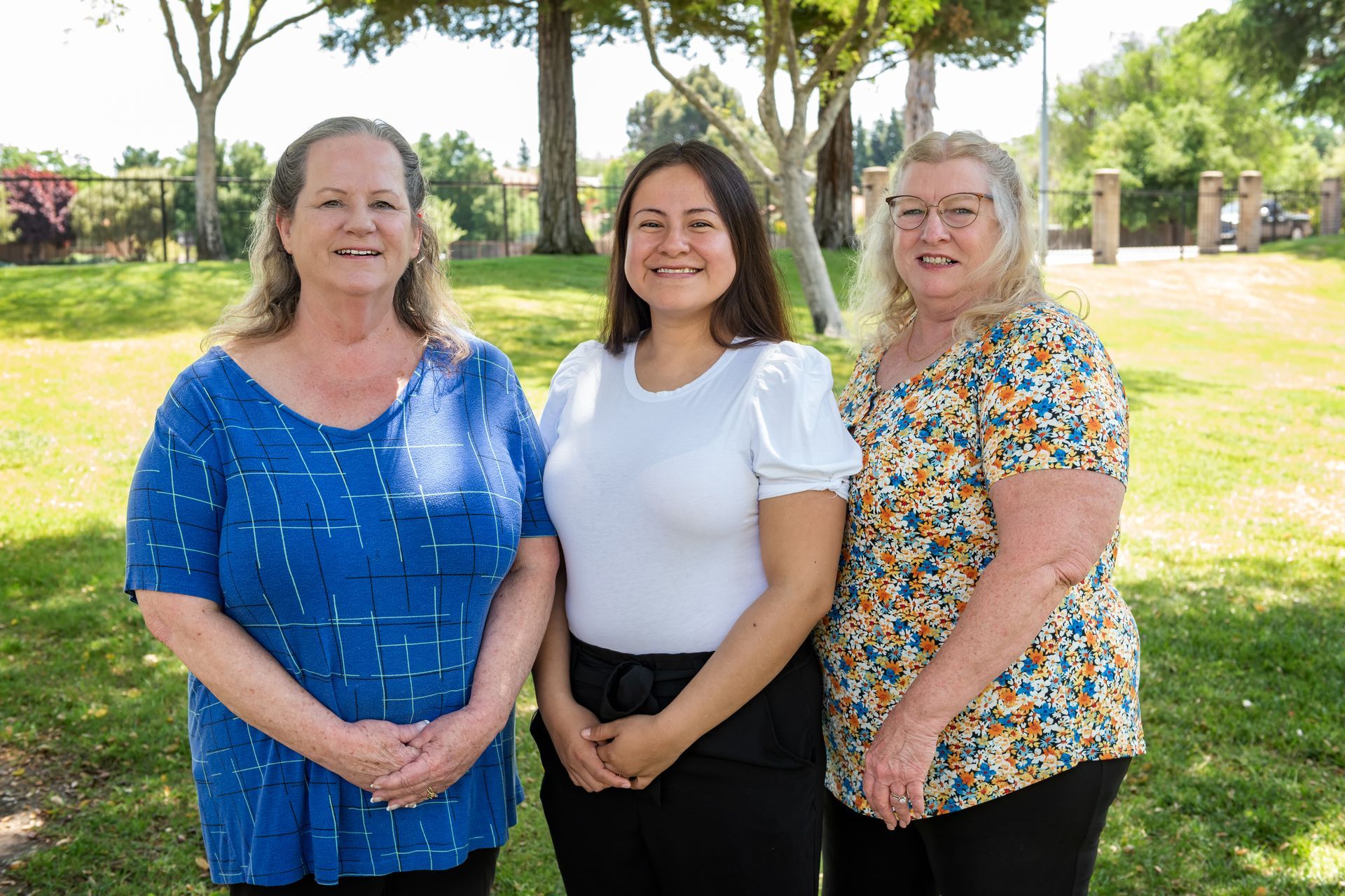 Three women smiling outdoors, standing in a park. They are in front of greenery and trees on a sunny day.