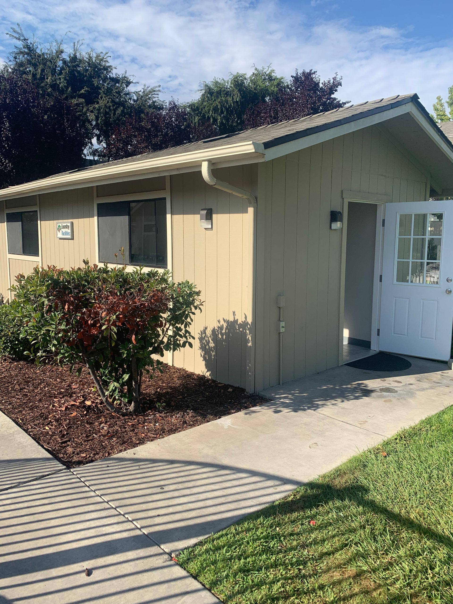 Beige building with open white door, sidewalk, grass, and a shrub.