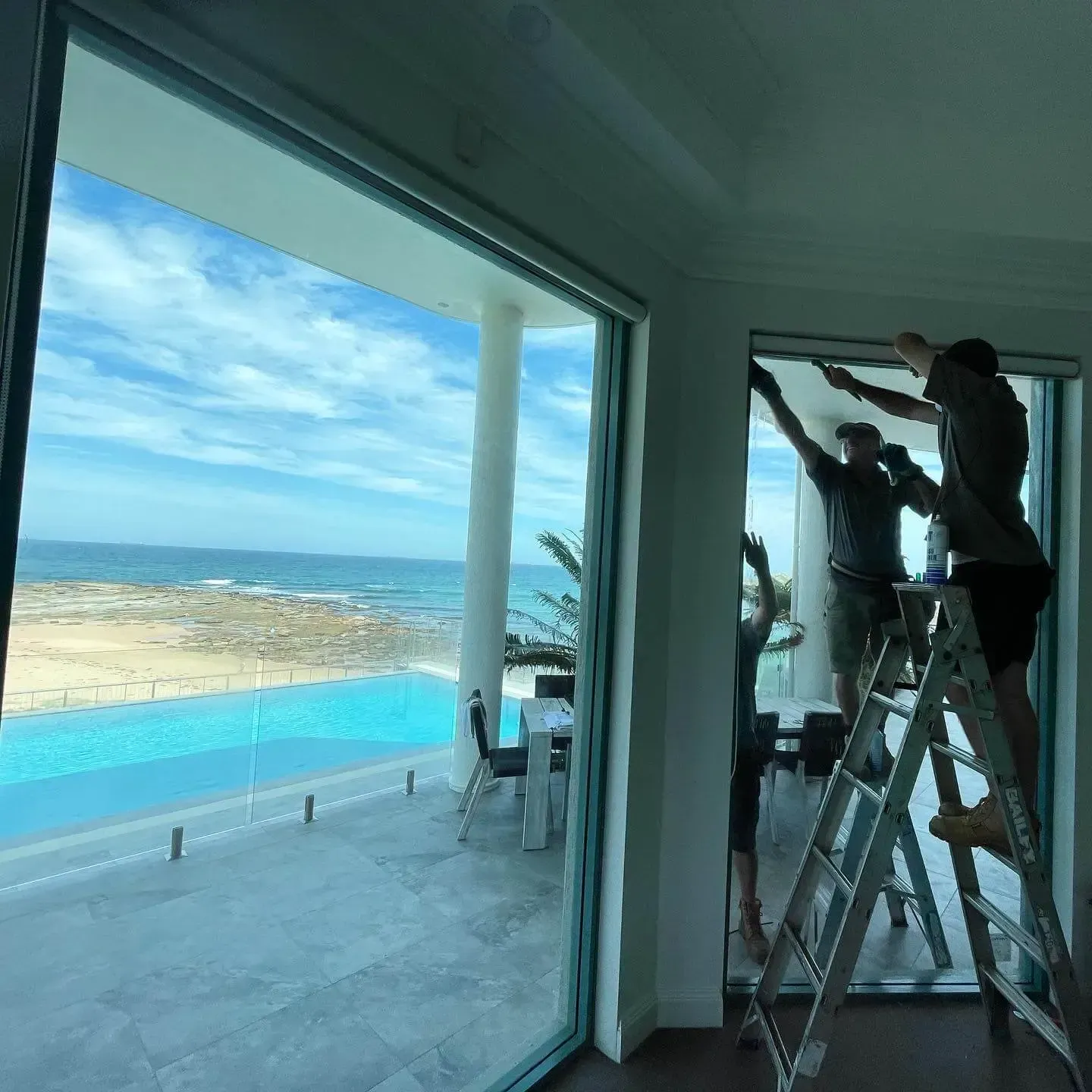 Two Men Are Standing on A Ladder in Front of A Window with A View of The Pool — Tuggerah Lakes Glass In Tumbi Umbi, NSW