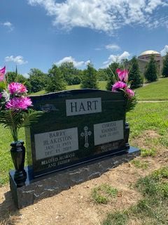 Headstone for Hart family, black granite, pink flowers, green grass, blue sky, domed building in background.