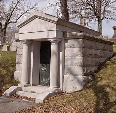 Stone mausoleum with columns at a cemetery. Green door and steps in front.