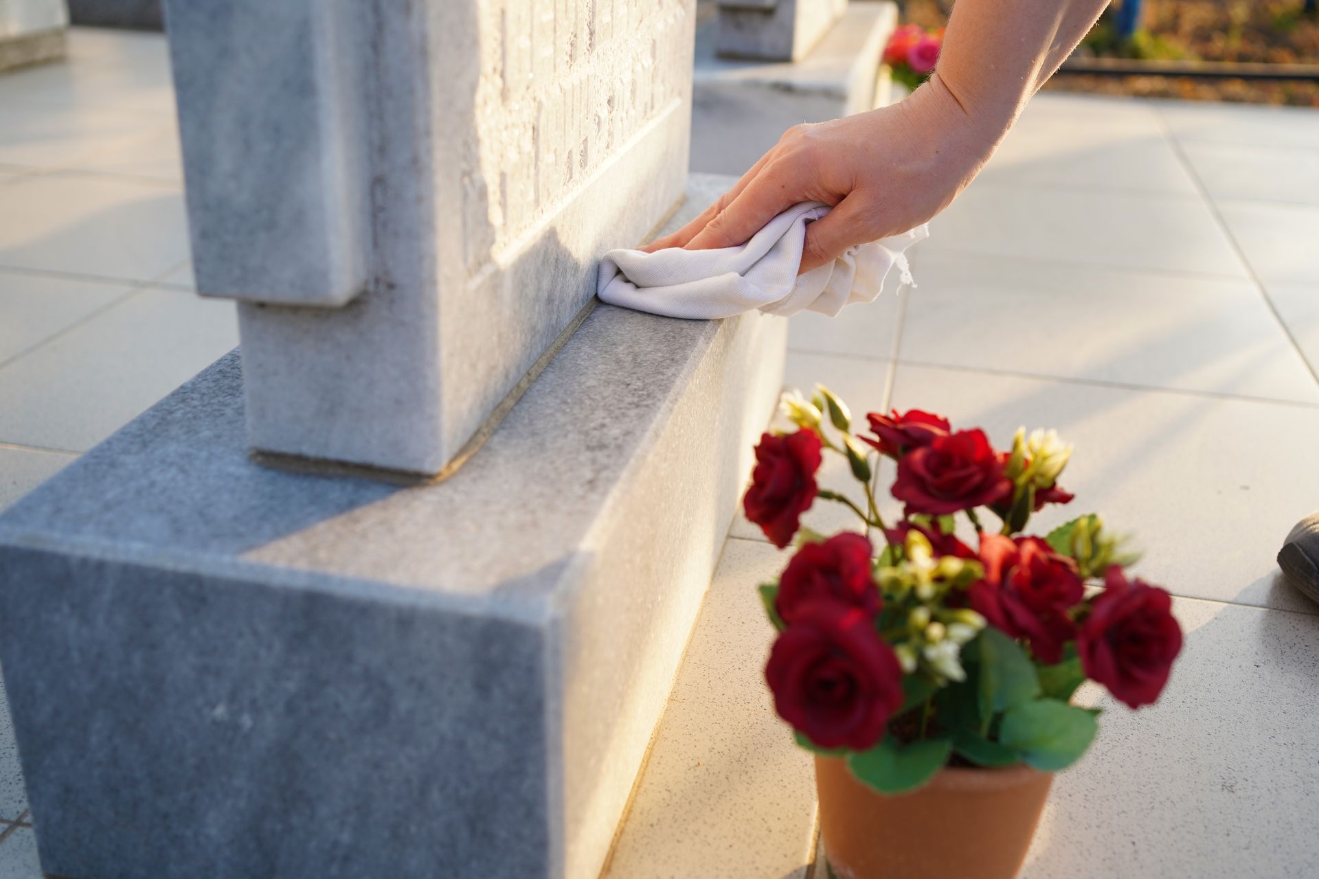 Person cleaning a marble headstone with a white cloth, near a pot of red roses.
