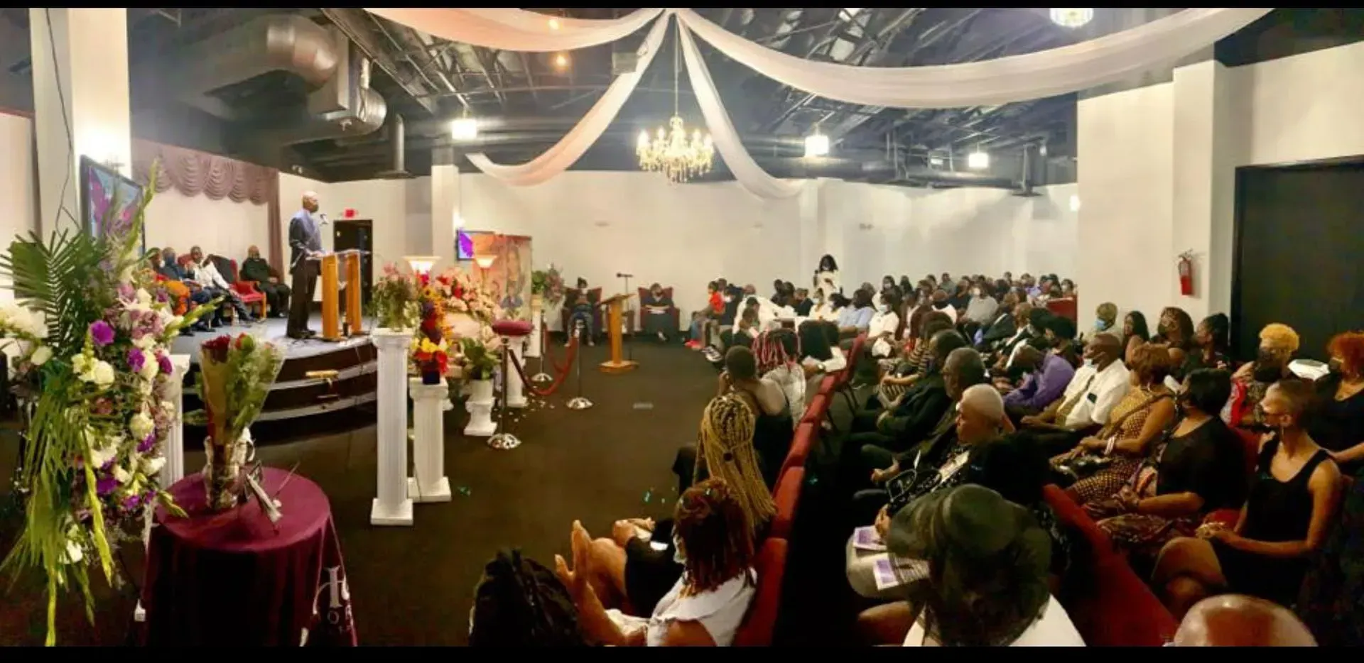Decorated hall with seated audience at a formal indoor event, flowers and draped ceiling lights visible