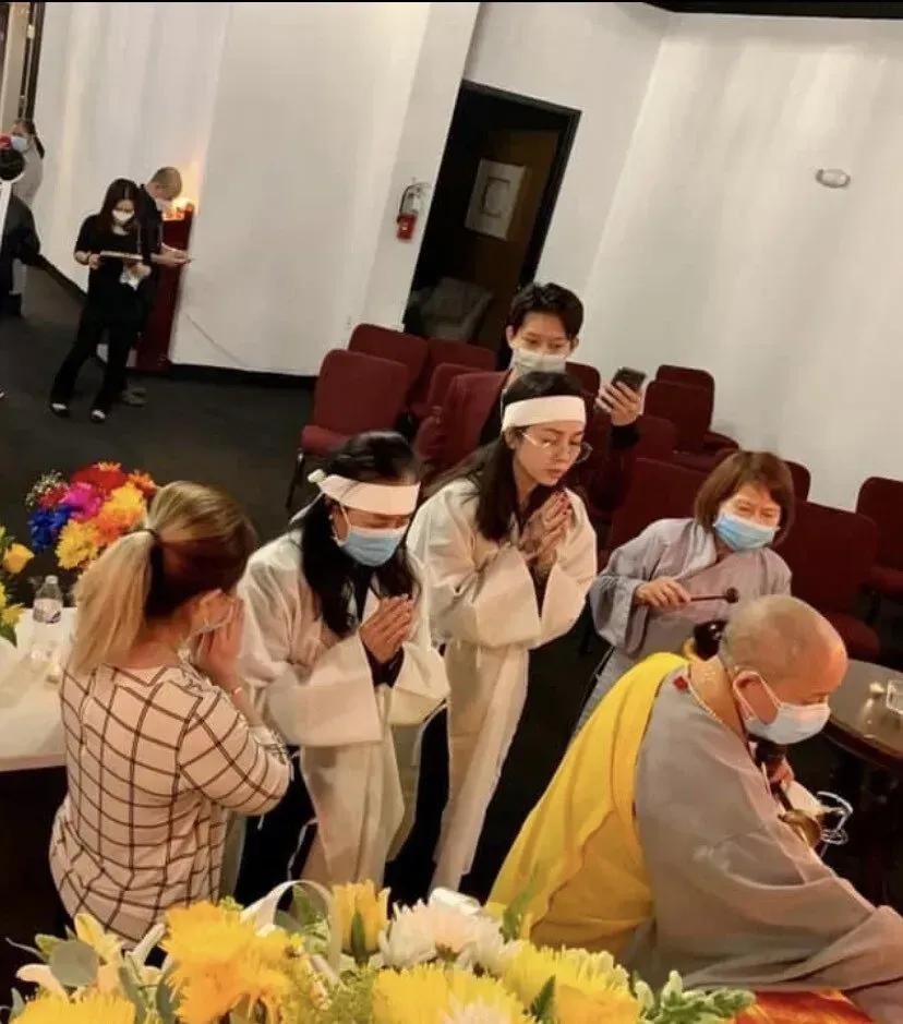 People in white robes and face masks stand in a quiet room beside a seated monk in yellow robes.