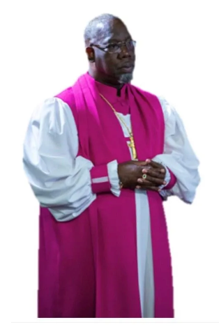 Man in a magenta and white clerical robe, standing with hands clasped against a white background