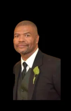Man in a black suit with a white shirt, black tie, and green boutonniere against a black background