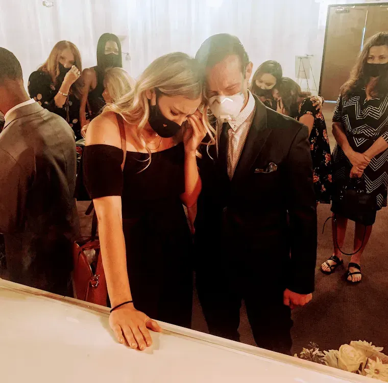 Masked couple in formal attire leaning over a white casket at a funeral service