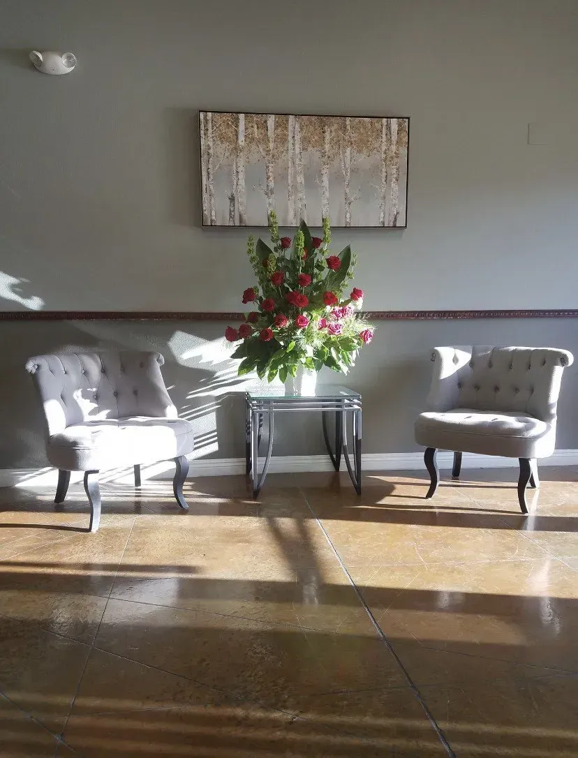 Sunlit lobby with two white armchairs, a small table with flowers, and wall art on a gray wall
