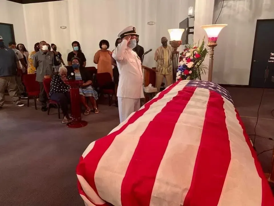 Draped casket with red and white stripes at a memorial service, mourners seated in the background