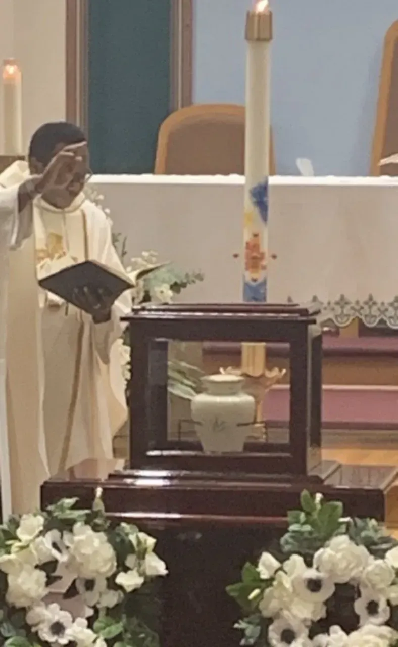 Clergyperson in white robes reading at a church altar beside a framed floral display