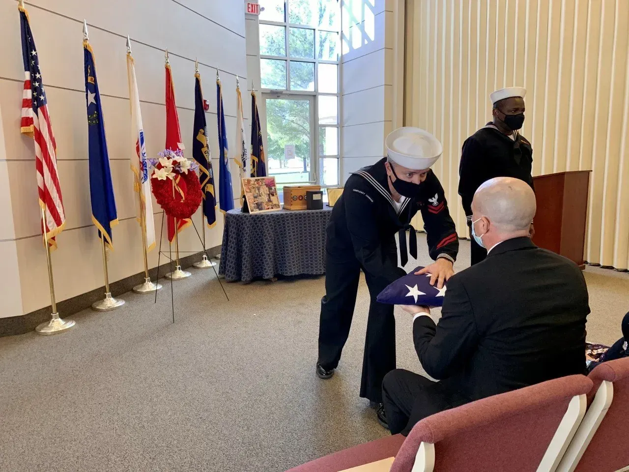 Ceremony in a hall as a masked person presents a folded flag to a seated man near flags and a podium