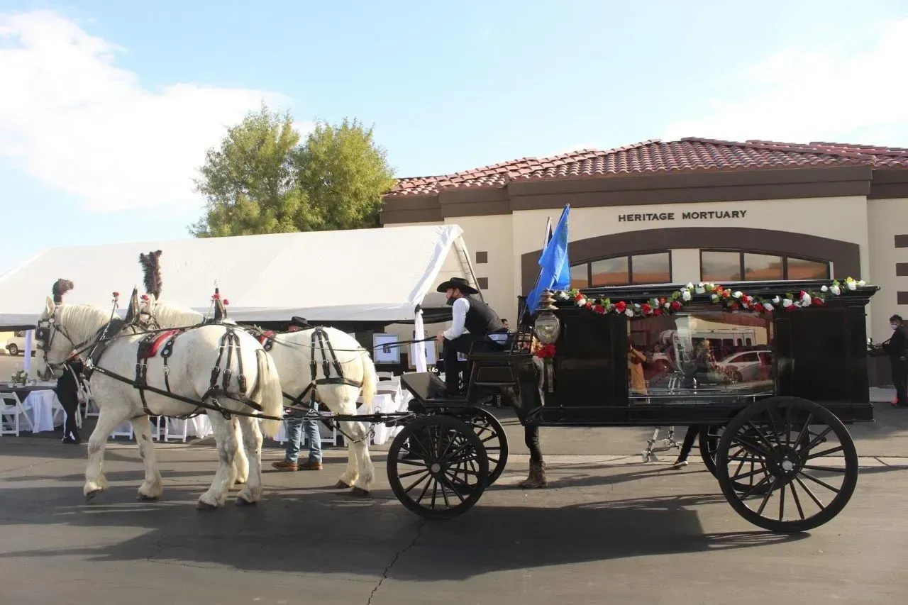 Horse-drawn black carriage decorated with flowers, parked outdoors beside white horses and a white tent.