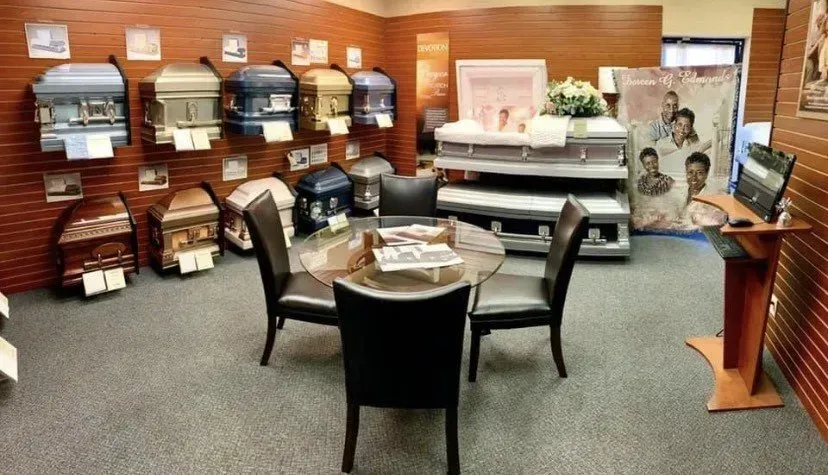 Showroom with wooden wall displays, upholstered chairs, a round table, and a coffin in the back