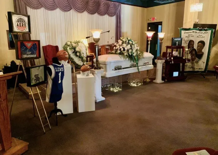 Memorial display with white casket, floral arrangements, framed photos, and blue jersey in a decorated room