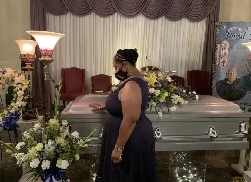 Woman standing beside a gray casket in a funeral home with floral arrangements and a memorial backdrop.
