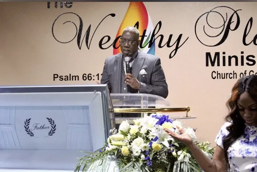 Speaker at church podium with floral arrangement, in front of a “Wealthy Place Ministry” banner.