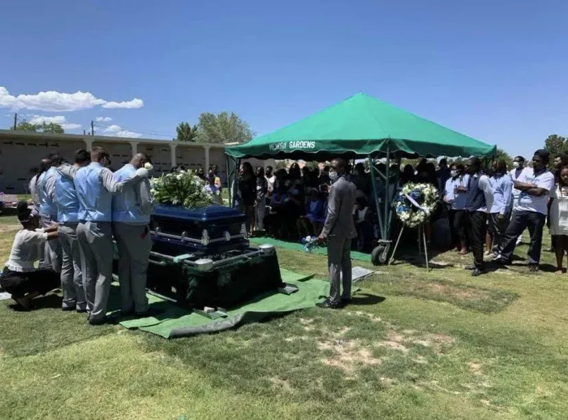 Outdoor gathering under a green canopy with people standing around a casket on a grassy lawn.
