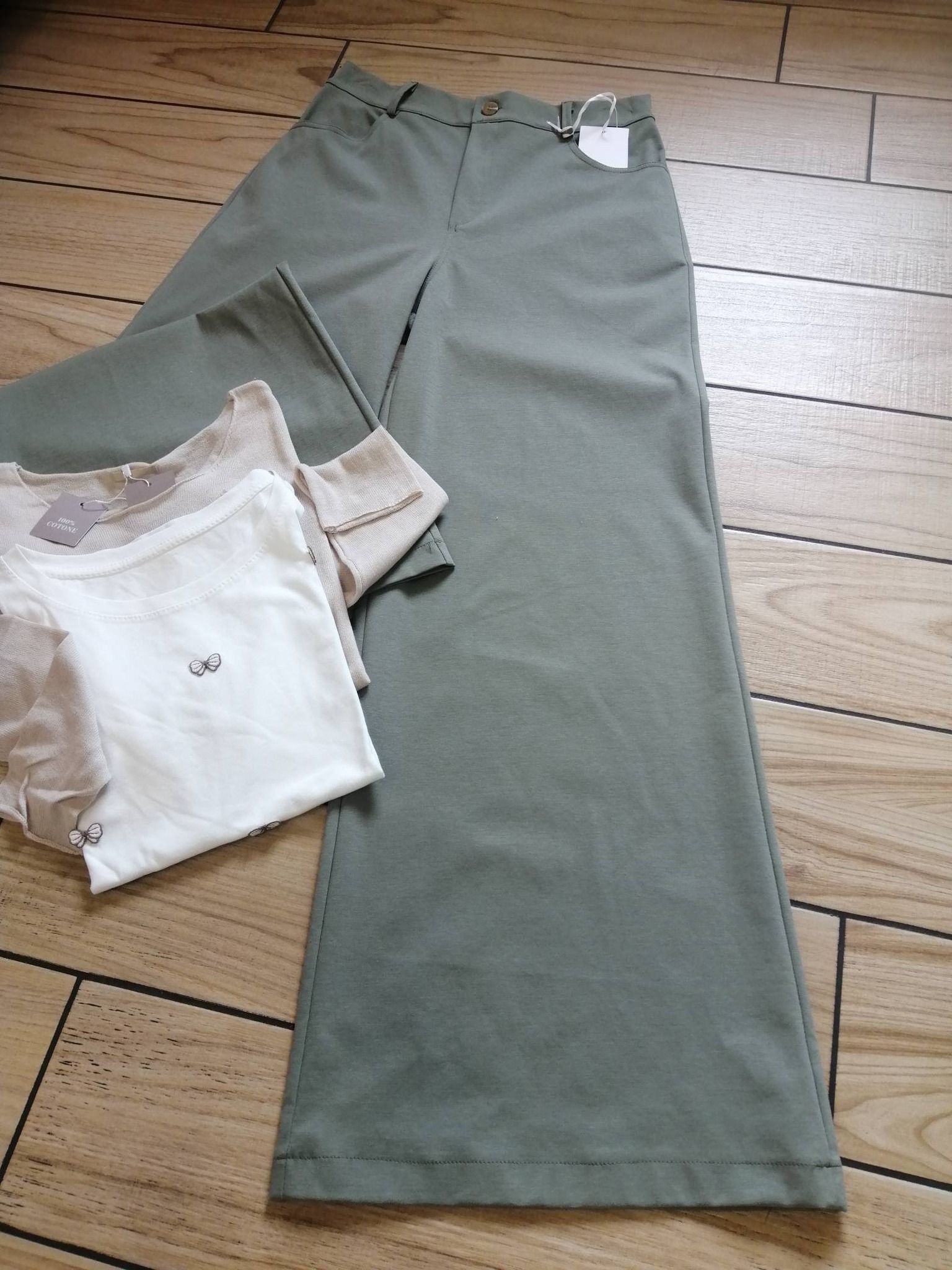 Pantaloni a gamba larga verde salvia a vita alta piegati accanto a una t-shirt bianca con stampa grafica su un pavimento di legno.