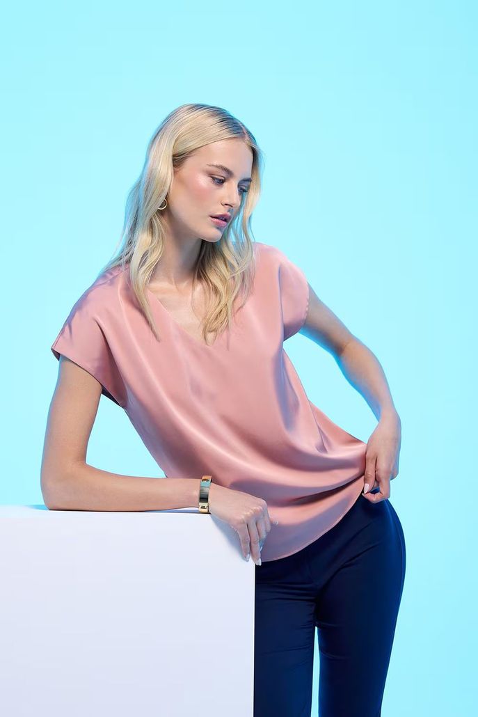 Una persona con i capelli biondi, che indossa una maglietta a maniche corte rosa cipria e pantaloni scuri, si appoggia a un blocco bianco contro una luce blu.
