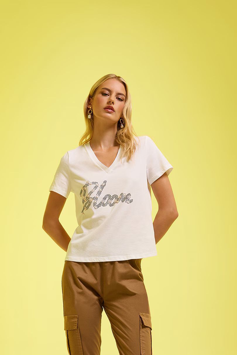 Una persona è in piedi davanti a uno sfondo giallo, indossa una maglietta bianca con scollo a V e scritta nera e pantaloni cargo color cachi.
