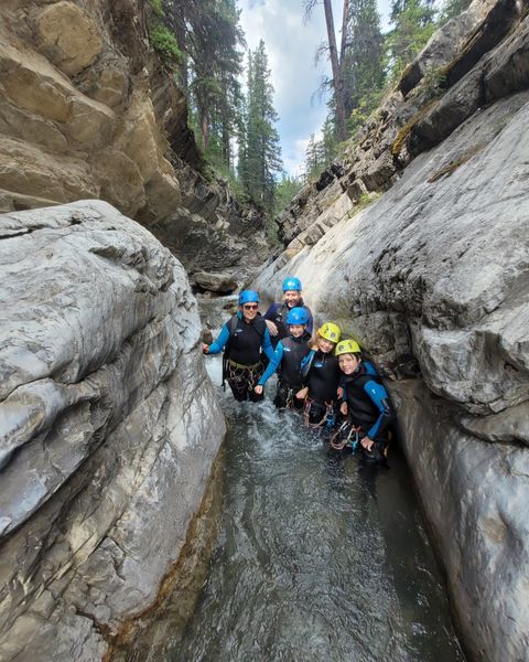 Five people in wetsuits stand in a narrow canyon stream. They wear helmets, looking forward.