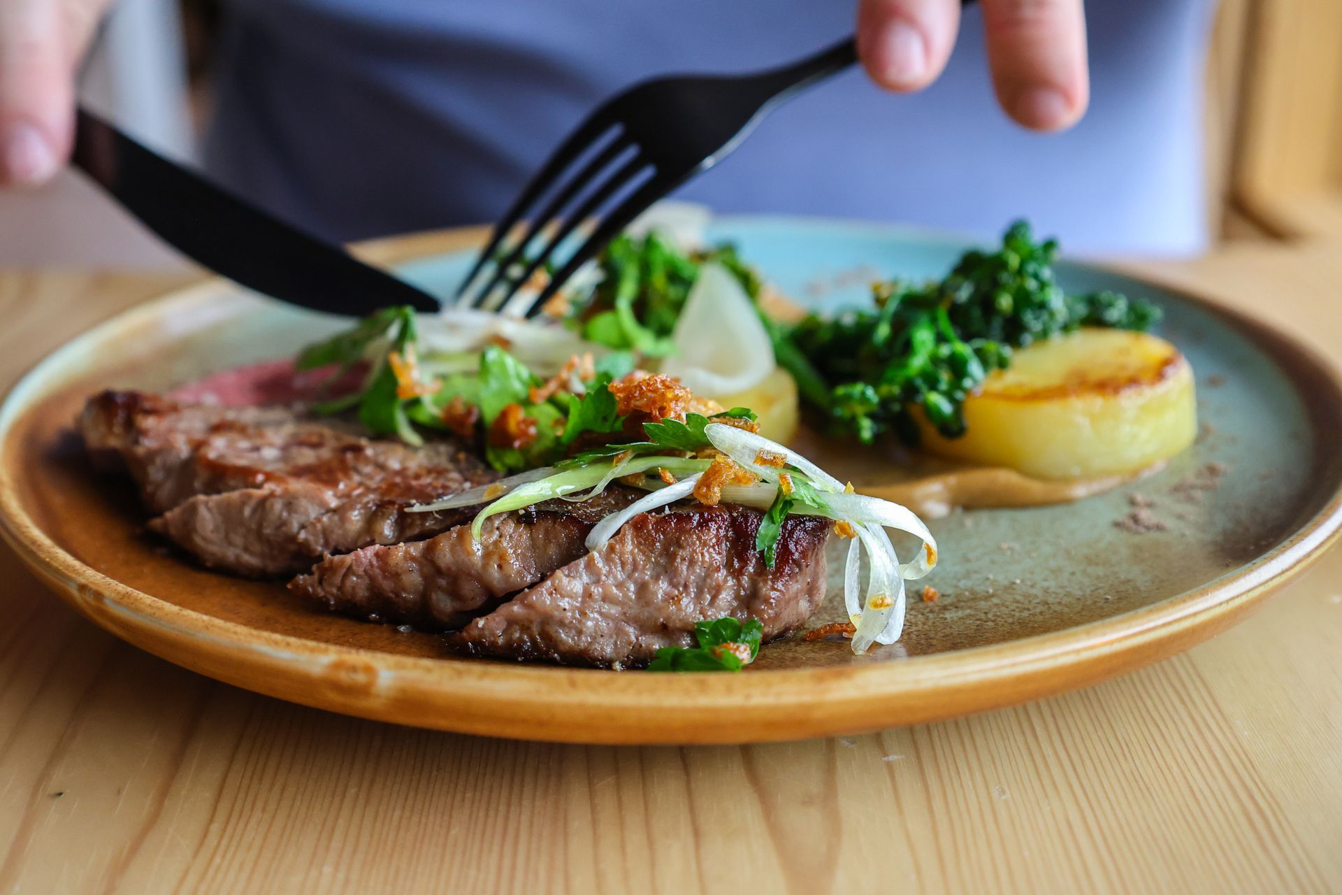 Person cutting steak on a plate with greens and potato.