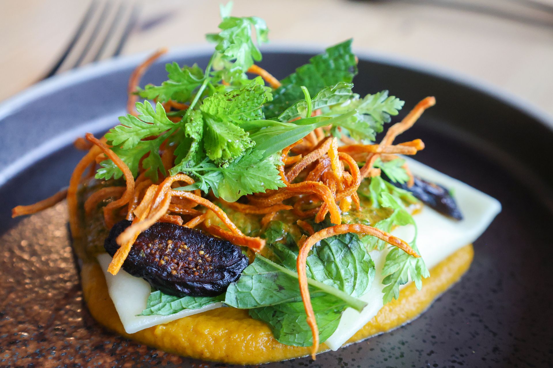Savory dish: yellow puree base, topped with white cheese, figs, herbs, and crispy carrot strings on a dark plate.