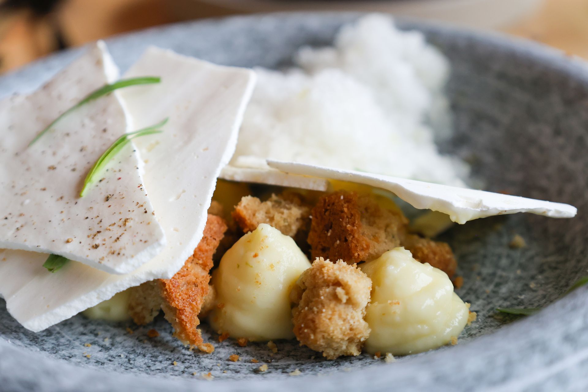 Close-up of a deconstructed dessert in a stone bowl. Components include white wafers, cream puffs, and crumbs.