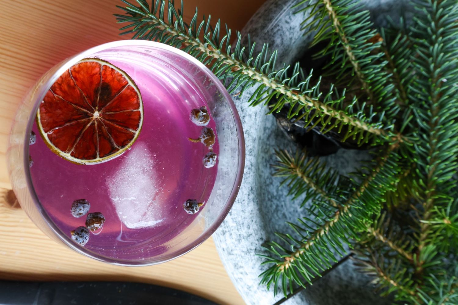 Cocktail with a dried citrus garnish, ice, and dark berries, beside a sprig of evergreen.