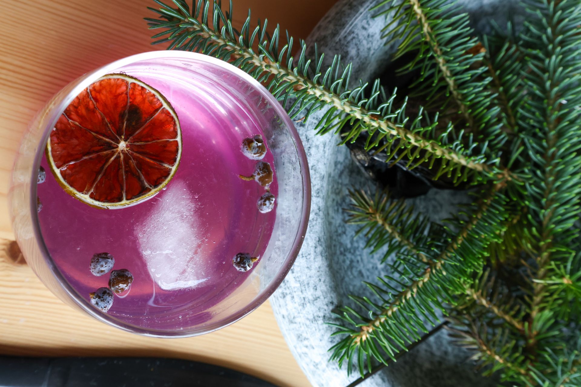 Cocktail with a dried citrus garnish, ice, and dark berries, beside a sprig of evergreen.