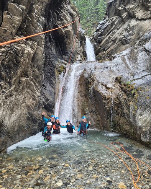 Group of people in wetsuits stands in a cold, clear stream beneath a waterfall.