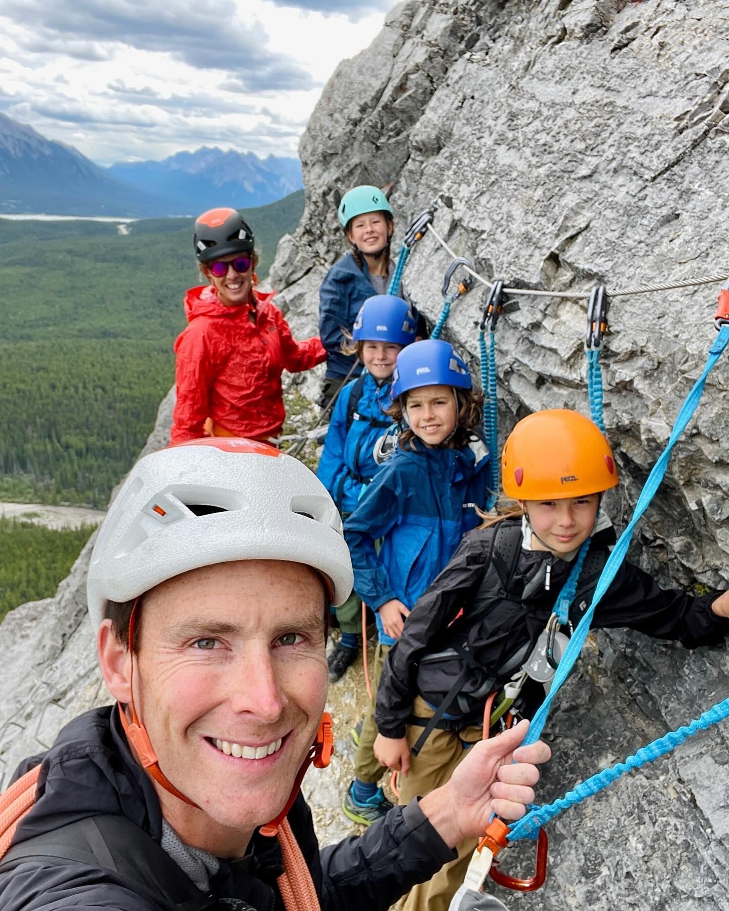 A man with a helmet smiles for a selfie, leading a group of children on a via ferrata climb in mountains.