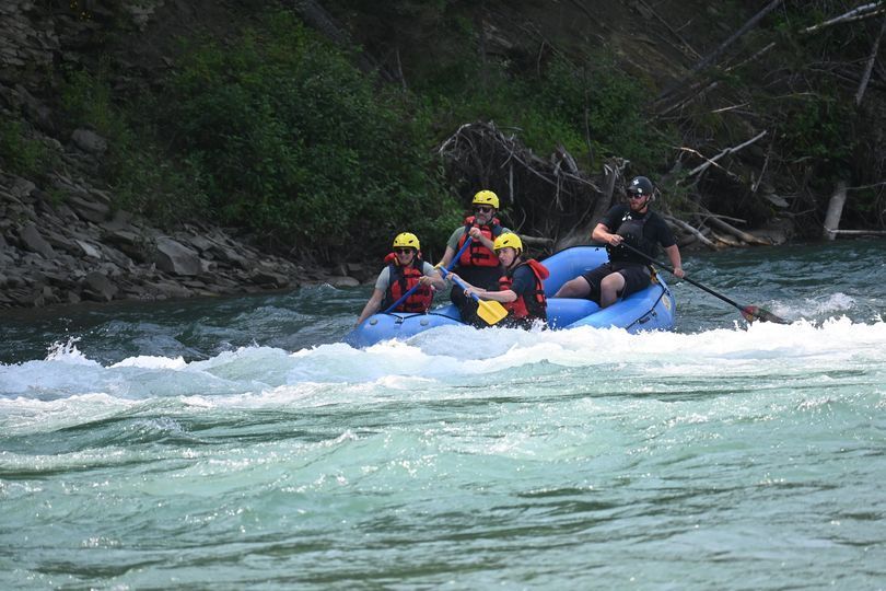 Raft with five people navigating a whitewater river. People wear helmets, paddling. Water is choppy, green.