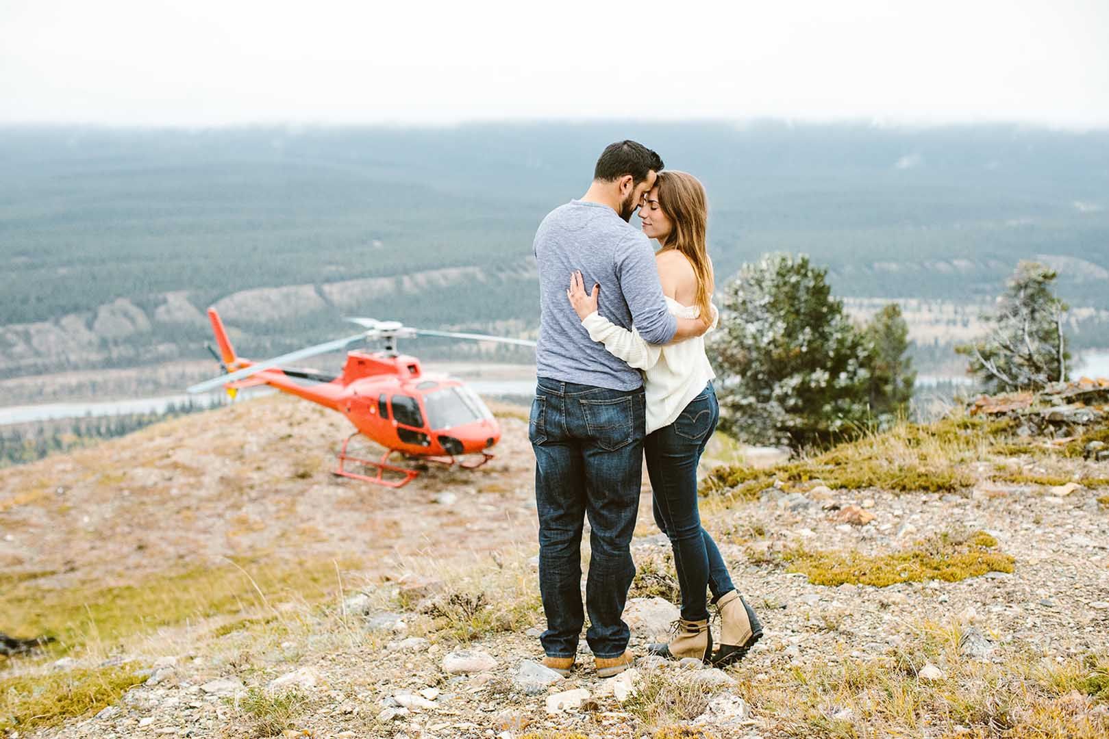 Couple embracing near a red helicopter on a hilltop overlooking a forest and river.
